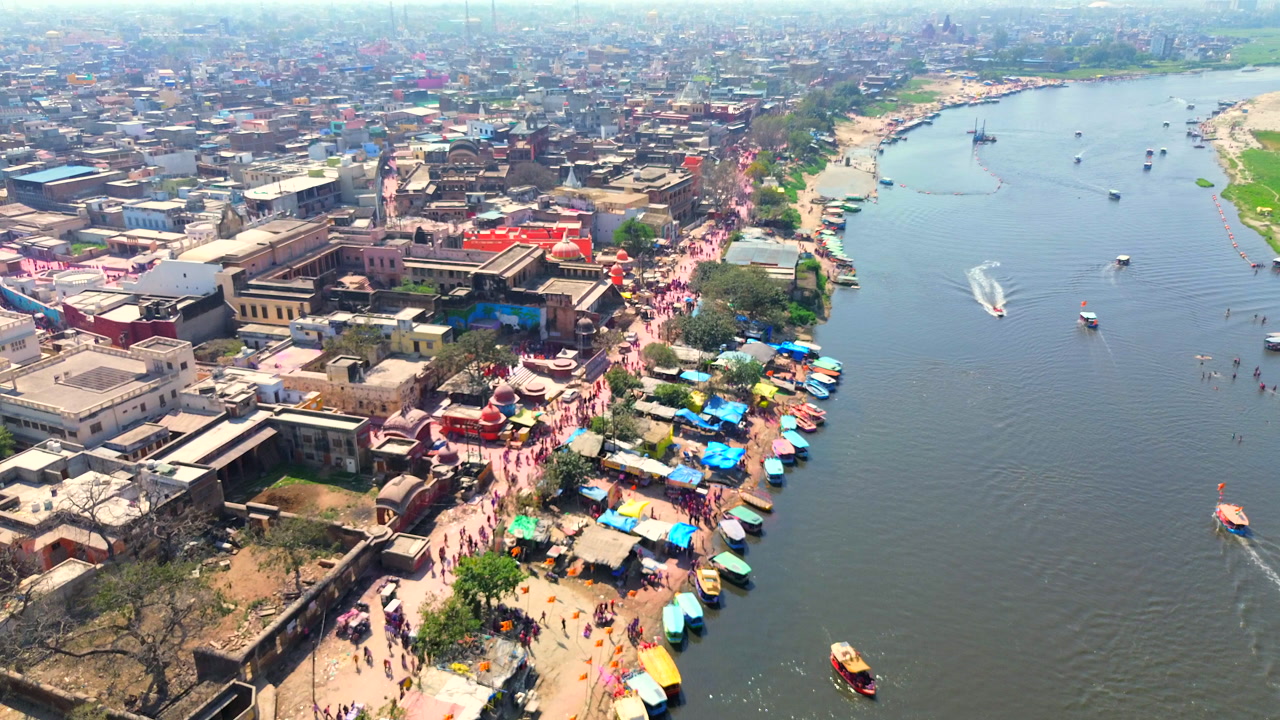 A dynamic drone tracking shot along the Yamuna River bank in Vrindavan, capturing people walking and celebrating Holi with traditional boats on the water and the city in the background.