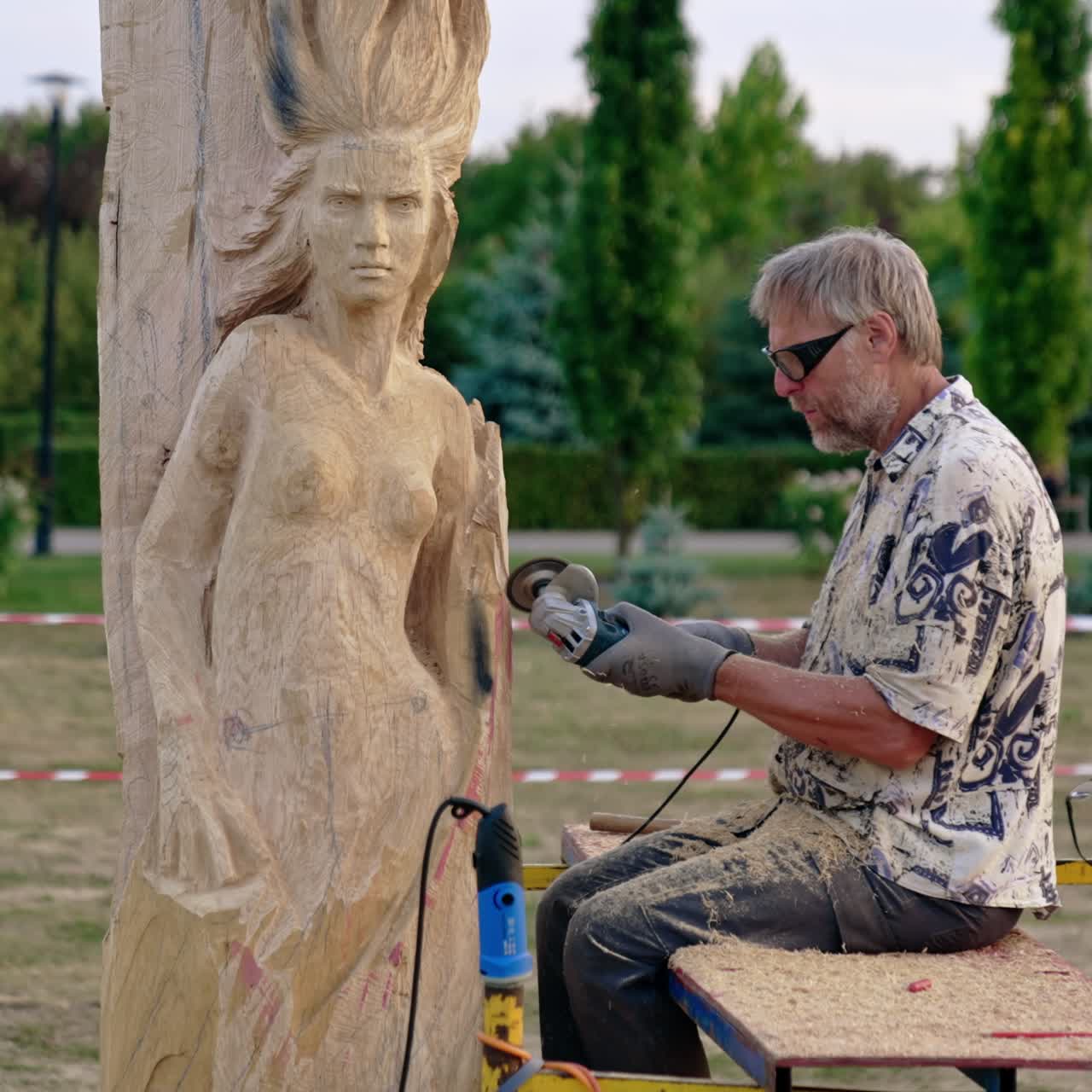 Craftsman sits outdoors working with an electric grinding instrument. Artist creating a wooden statue of a woman