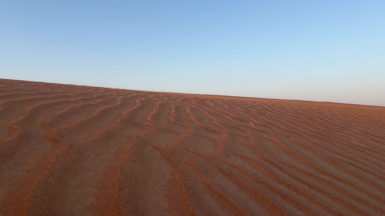 A close up of the desert and its red sand as wind is blowing particles of dust with a blue sky background.