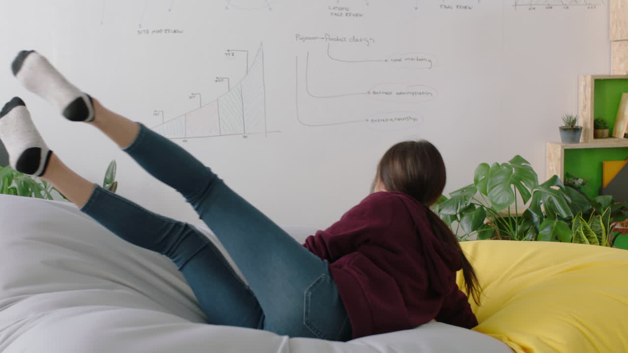 행복한 백인 여성 학생이 다채로운 베개에 뛰어들고 여자가 휴식을 취하고 트렌디한 사무실 작업장에서 점심 휴식을 즐기고 있습니다.