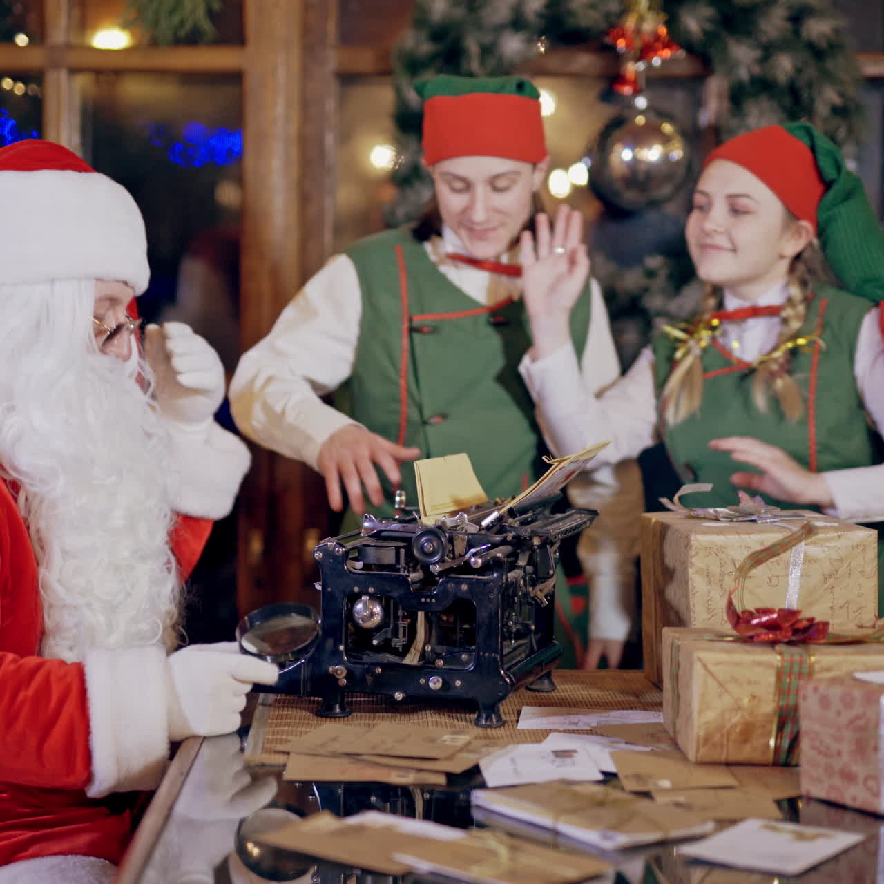 Santa Claus is sitting at the table and two joyful elves dancing on the Christmas background indoors. Many presents, letters on the table and Santa is printing on the old typewriter. Elves in green costumes and Santa prepare for the Christmas holidays.