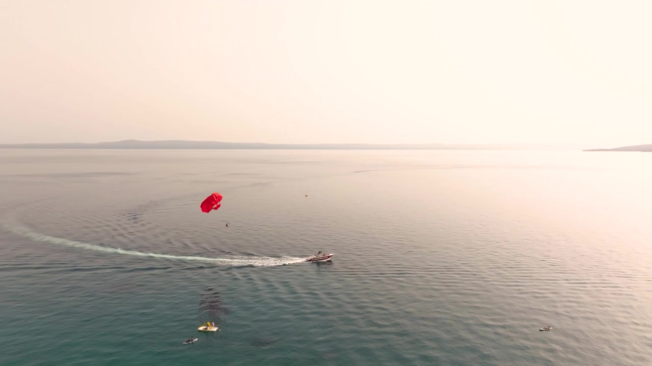 Red parasail and tow boat arc over quiet open water as smaller craft float nearby on a soft hazy day.