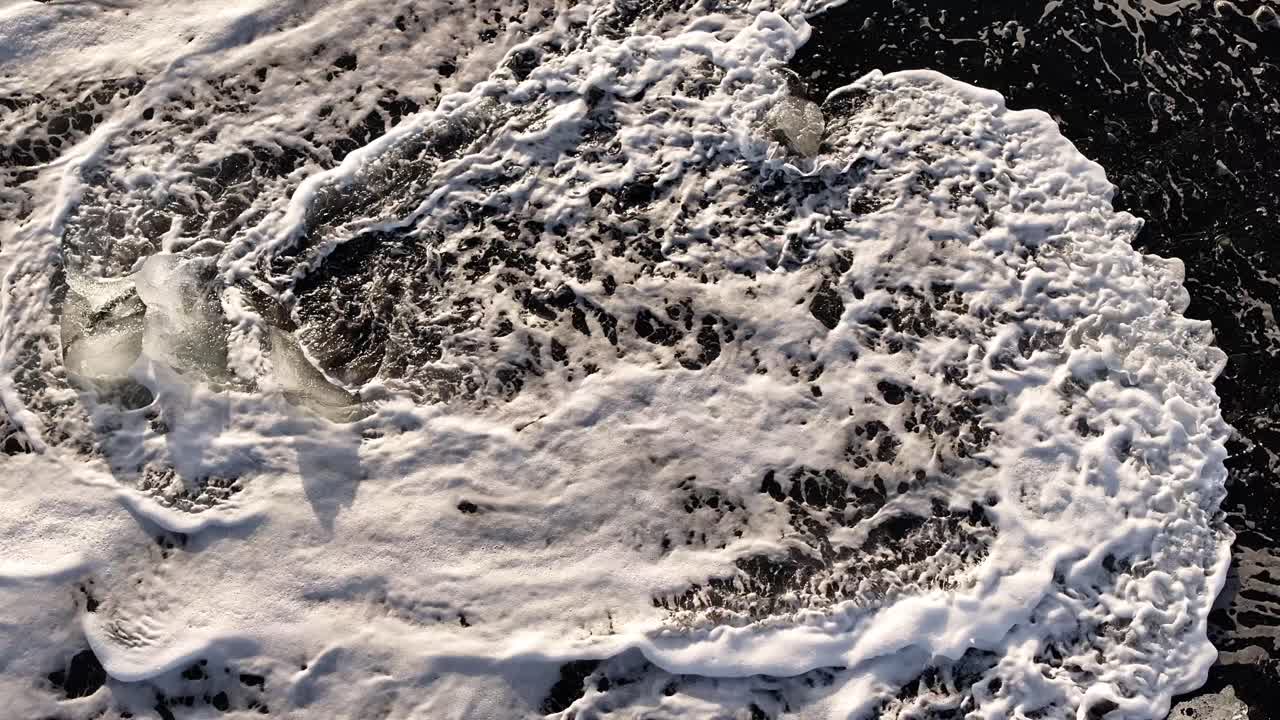 Overhead view of foamy ocean waves swirling over dark coastal waters in rhythmic motion. Icelandic coast