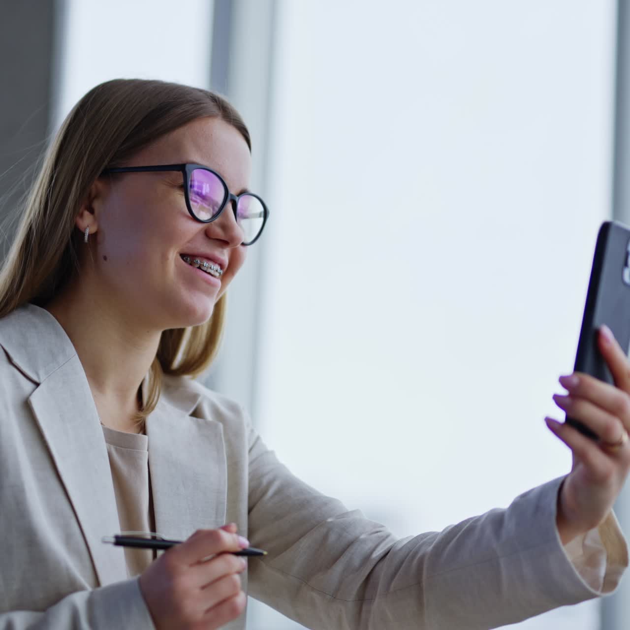 Positive young long-haired woman in glasses having online chat with client. Lady is talking and smiling while holding phone in front of her