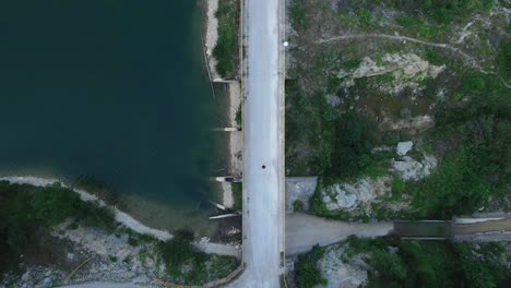 Vista-De-Pájaro-Del-Ojo-De-Arriba-Hacia-Abajo-Del-Dron-De-Cruzar-La-Carretera-Del-Puente-En-El-Paisaje-De-Montaña-De-La-Naturaleza-Con-Un-Lago-Claro-Y-Un-Bosque-Natural-De-Pinos