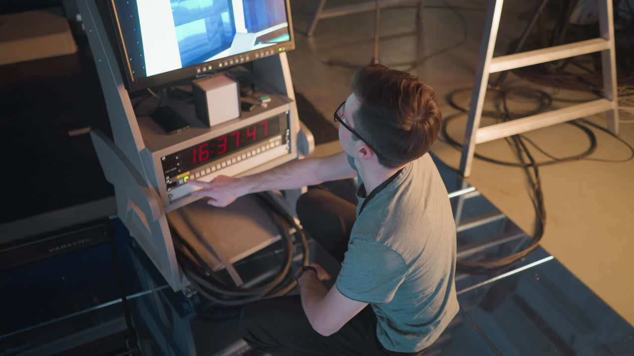 Person adjusting video equipment in TV studio, focused on monitor displaying broadcast setup. Timer on control panel shows time, studio lighting and cables visible in background