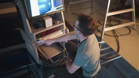 person adjusting video equipment in tv studio, focused on monitor displaying broadcast setup. timer on control panel shows time, studio lighting and cables visible in background