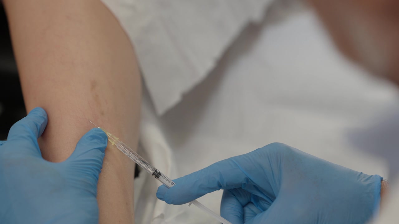 Gloved hands holding a small syringe against leg skin during a vein injection