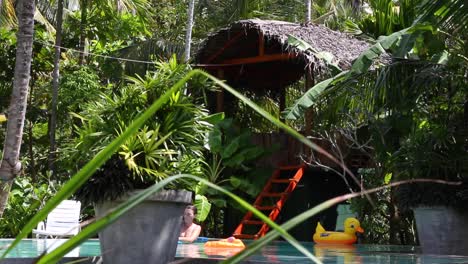 wide shot of a girl swimming in a pool at a luxurious tropical resort with a straw hut in the background