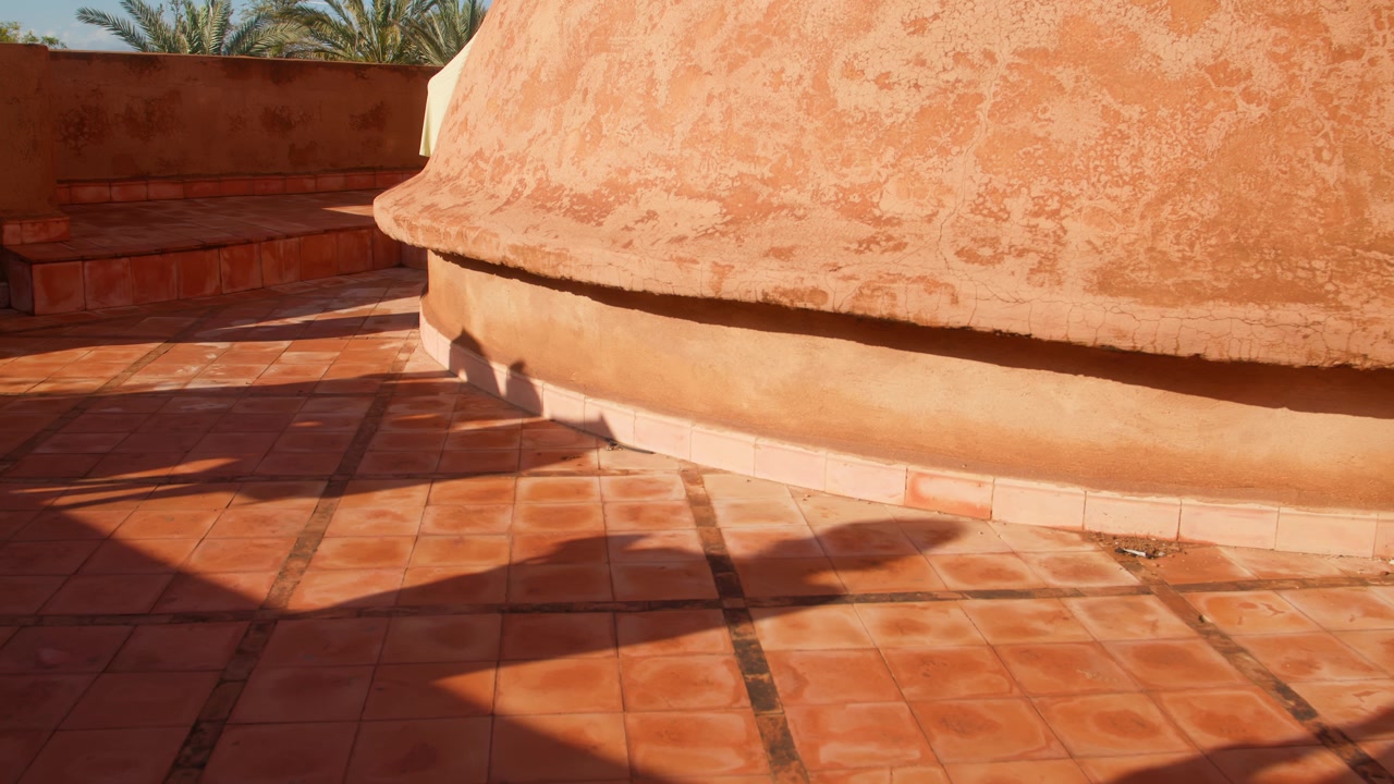 La calda luce del sole proietta ombre su piastrelle in terracotta e sulla parete curva di una villa a Marrakech.