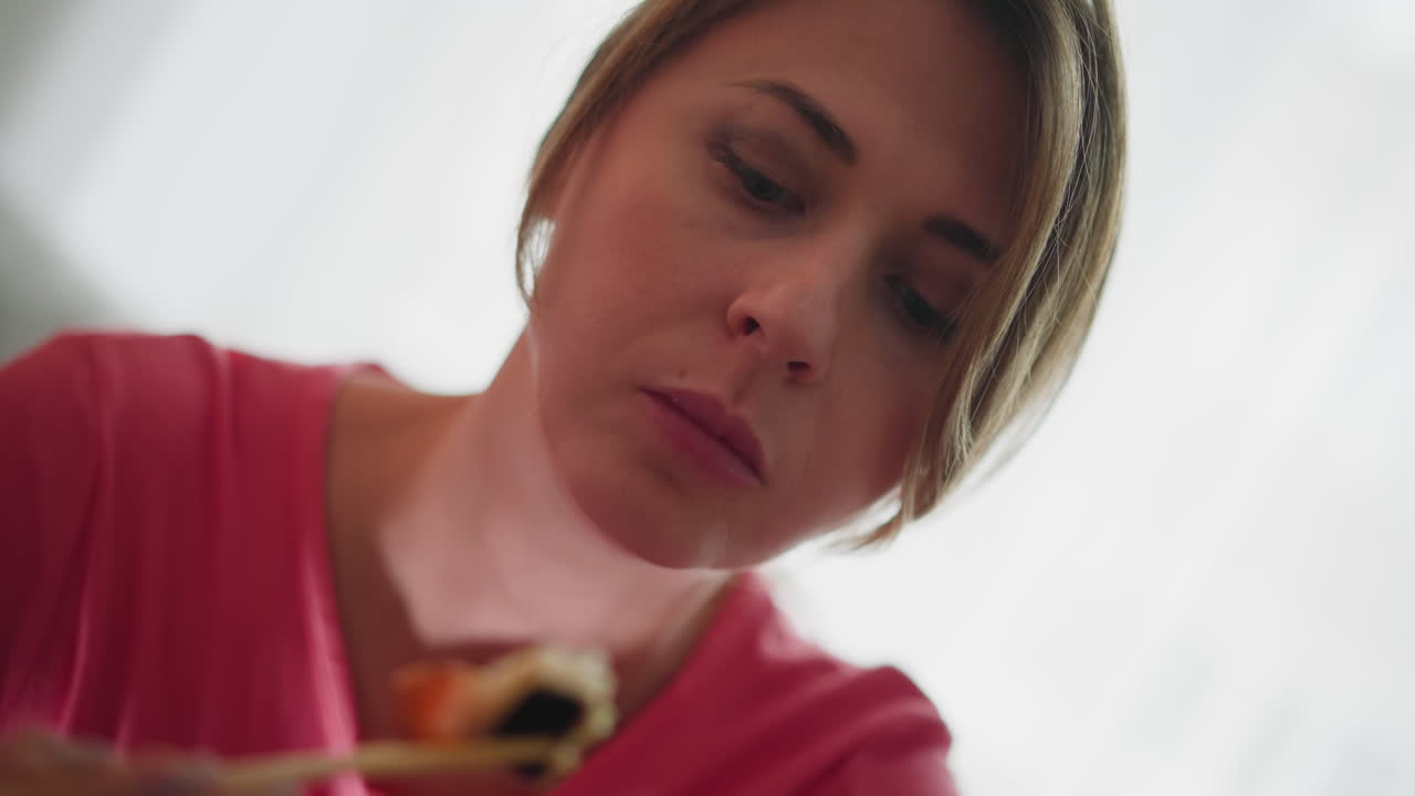 Girl in pink polo bending forward while eating sushi with chopsticks, showing focus and enjoyment as she savors each bite, captured in natural indoor light with soft blur in background