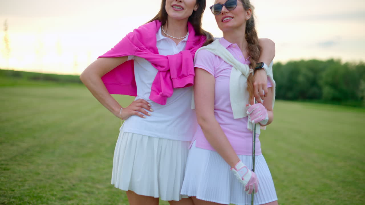 Two women in white and pink clothes, standing and holding golf clubs, on a grass field