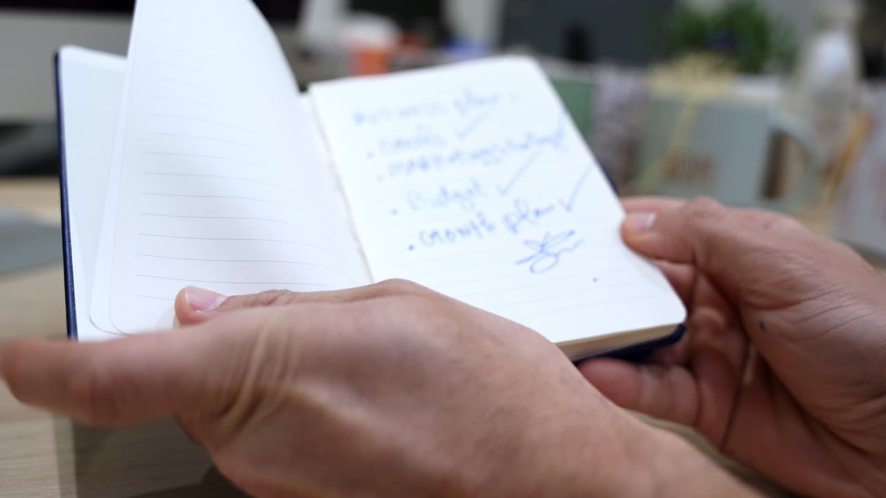 Close-up of hands holding an open notebook with a handwritten business plan checklist, including goals, marketing strategy, budget and growth plan in an indoor office setting