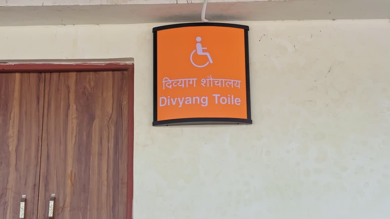 Tilt-up shot revealing an accessible toilet sign in India, featuring a wheelchair symbol and bilingual text, mounted above a doorway on a public facility wall