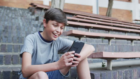 sitting on steps, boy using smartphone and smiling outside school building