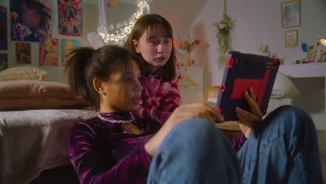 african american girl sits at floor, surfs internet using tablet. asian teen lies on bed and watches content with friend. happy diverse girls together spend leisure time at home. friends relationship.