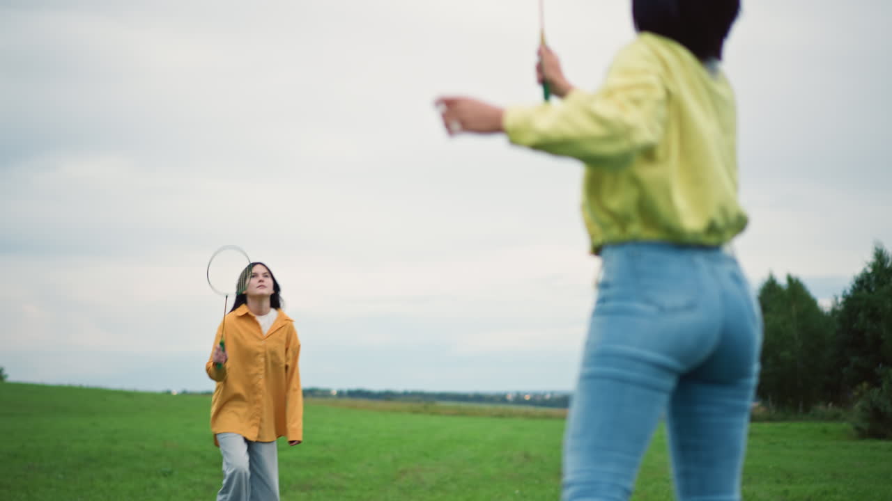 mulheres jogando badminton no campo: dupla animada treinando com raquete e peteca, usando jaqueta amarela casual e jeans. Movimento dinâmico e perseguição divertida em campo verde sob céu nublado, cena energética