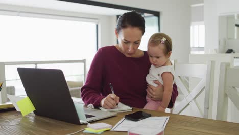 Kaukasische-Mutter-Hält-Ihr-Baby-Und-Macht-Sich-Notizen,-Während-Sie-Von-Zu-Hause-Aus-Arbeitet