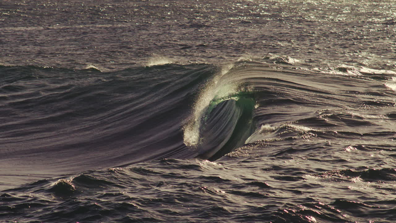 Sunset light hits large curling wave, golden tones glisten off textured crest in slow motion