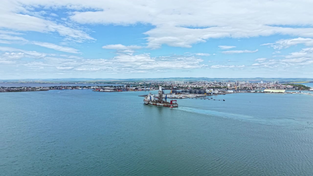 Промышленное дноуглубительное судно работает в воде у прибрежного города под облачным небом с видимой промышленностью на заднем плане