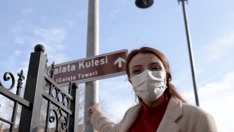 beautiful girl wearing protective medical mask and fashionable clothes shows sign of galata tower new normal lifestyle, travel concept