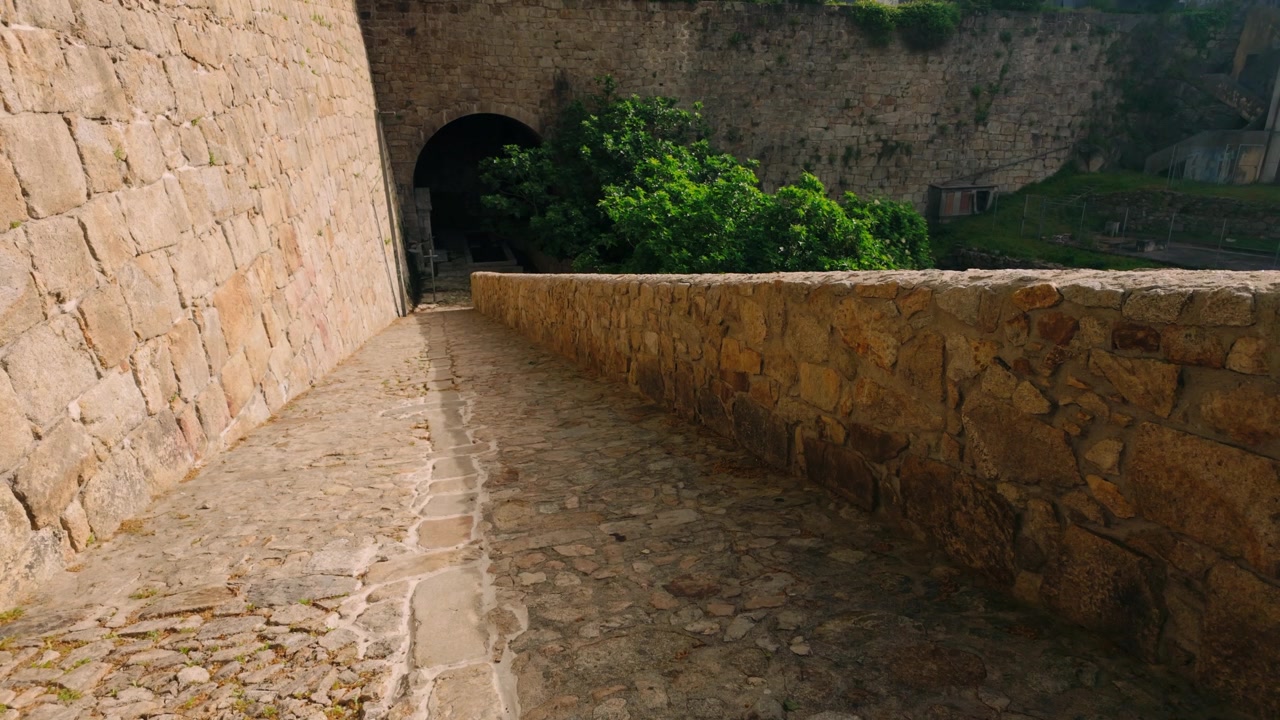 Historic cobblestone path winding alongside ancient stone walls, leading towards a dark, mysterious tunnel entrance. Lush green vegetation thrives along the weathered brickwork under bright natural...