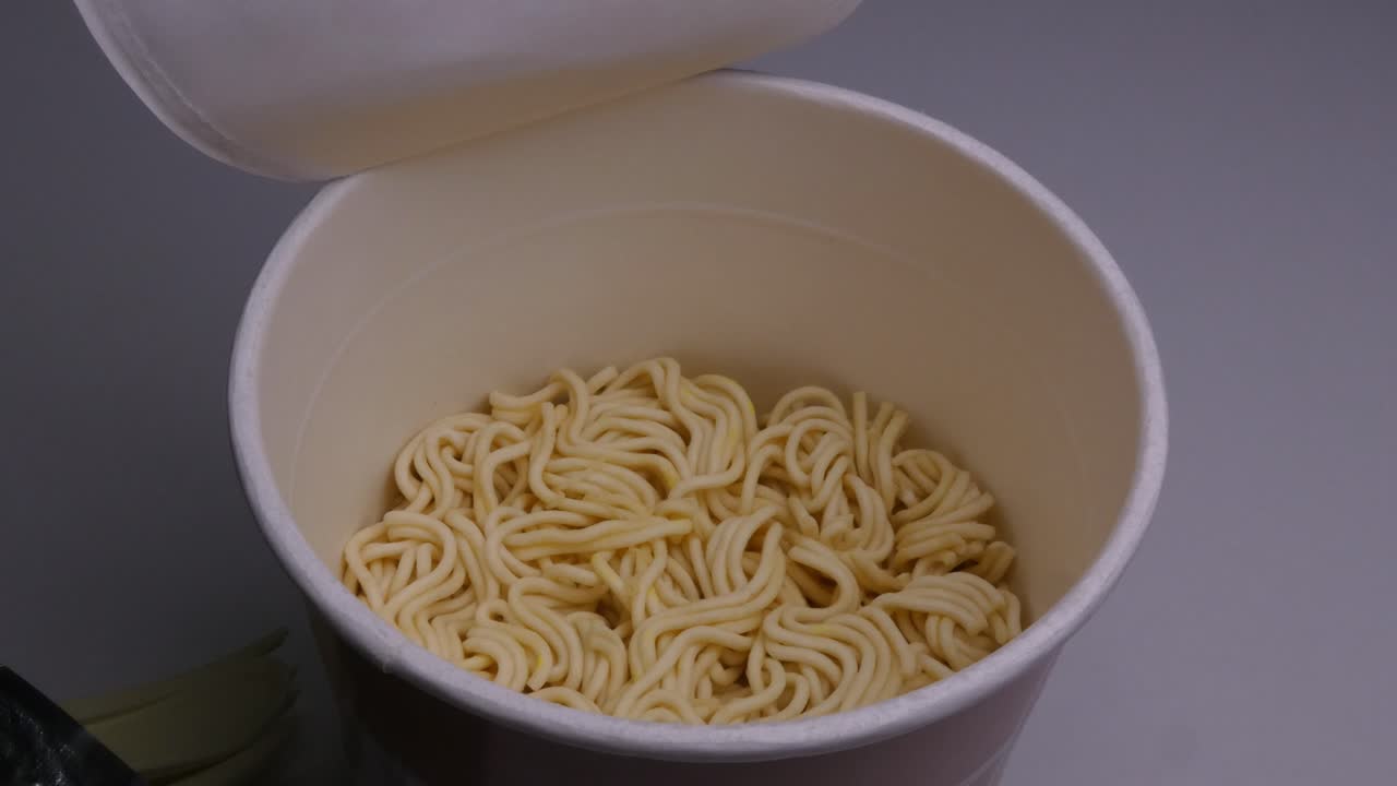 Un ramen istantaneo aperto e una mano che estrae da dentro una forchetta di plastica piegata e delle bustine di condimento, il tutto sopra i noodles essiccati
