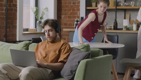 jongen die op een laptop werkt terwijl hij op de bank zit terwijl zijn kamergenoten de keuken schoonmaken