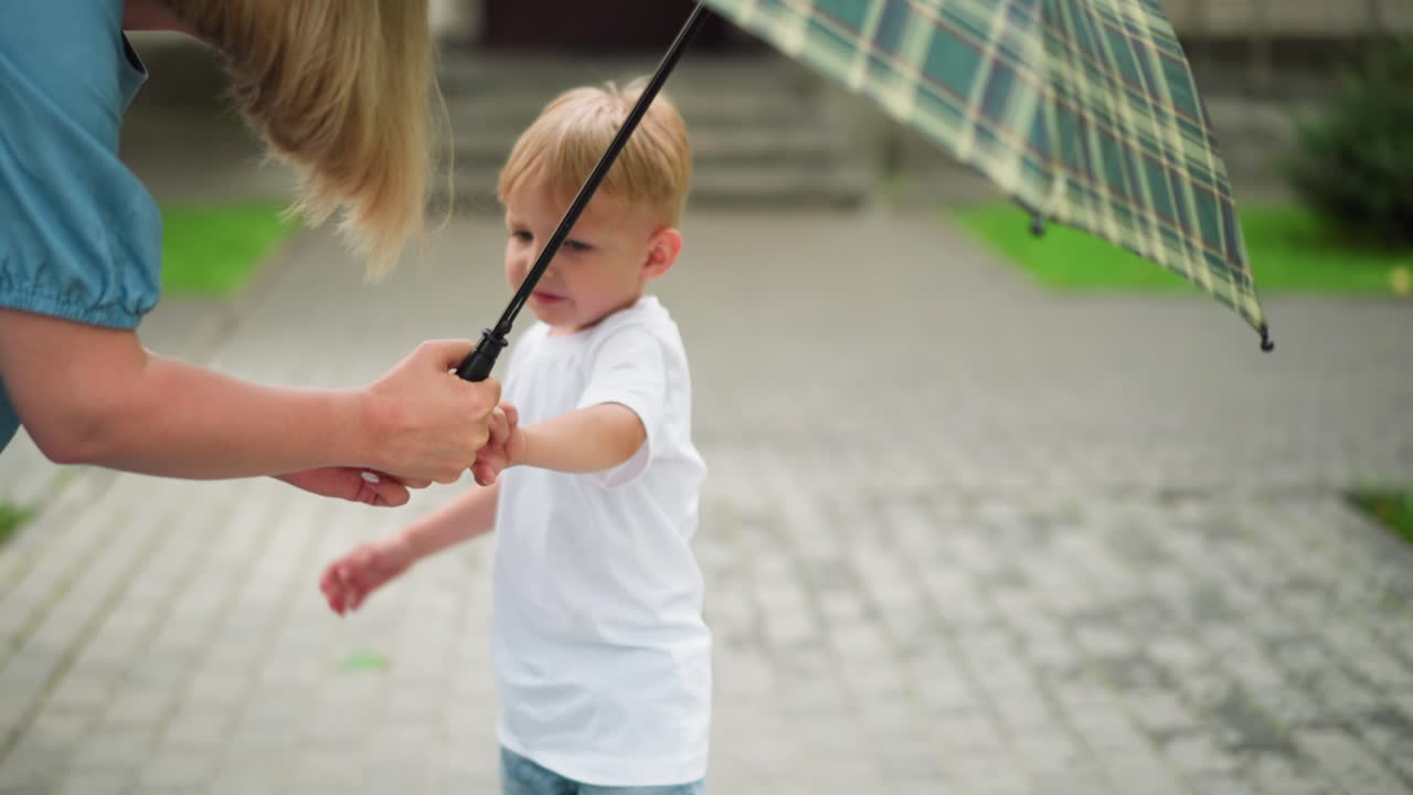 A little boy is seen trying to collect an umbrella from his mother, looking unhappy, with another child a little bit visible by the side