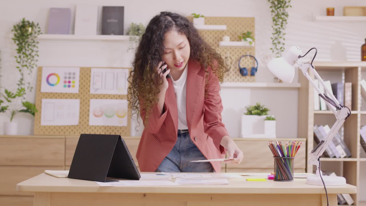 Asiatisk designer kvinde bruger sin smartphone til at kommunikere. Arbejder på projekter og dokumenter og arbejder hjemmefra ved et skrivebord med en bærbar computer.