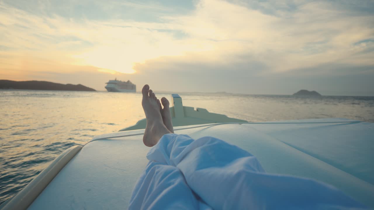 Point-of-view of a person relaxing on a boat deck during golden hour, calm sea and cruise ship in the distance