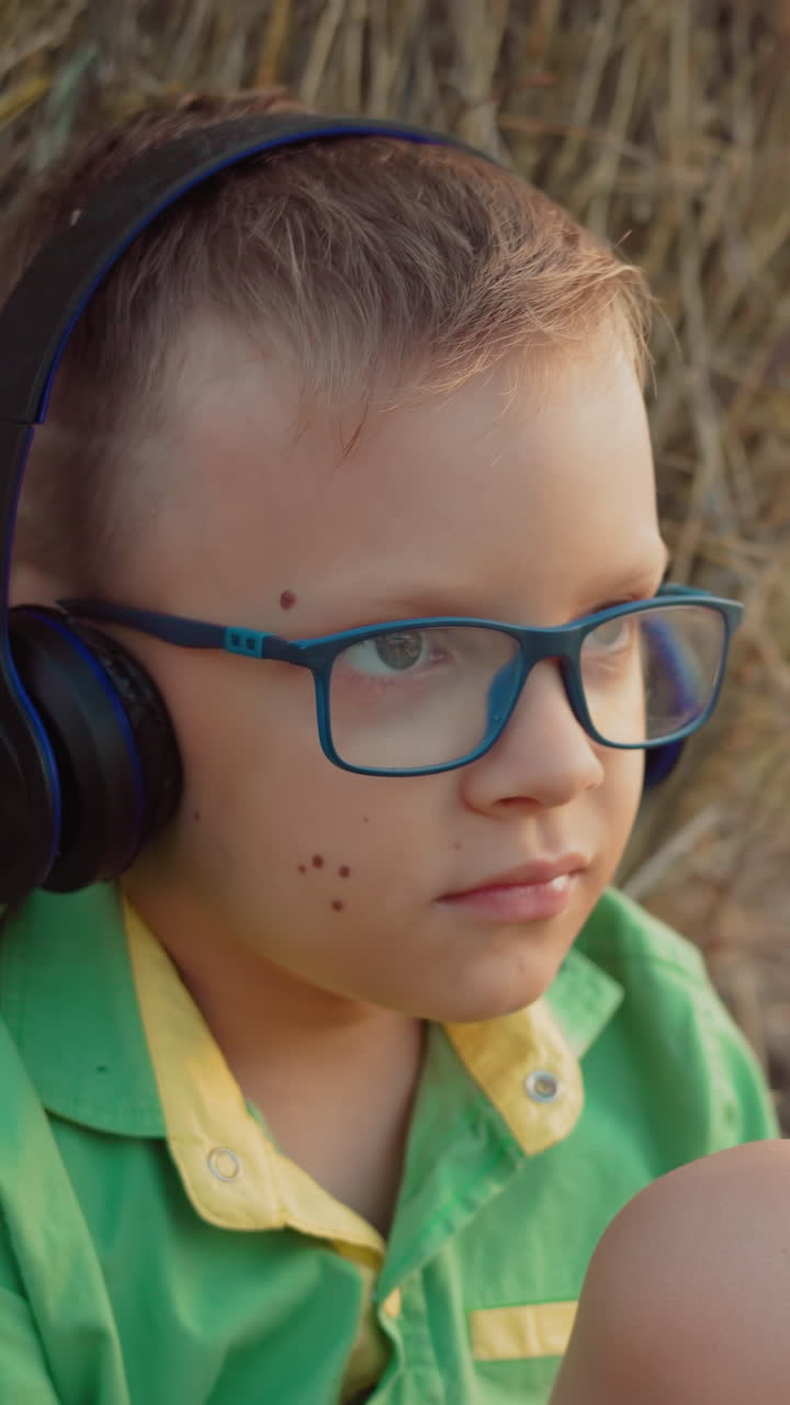 niño pequeño con auriculares y gafas profundamente centrado en escuchar música, sentado contra el heno en una vasta tierra de cultivo al atardecer, momento al aire libre sereno