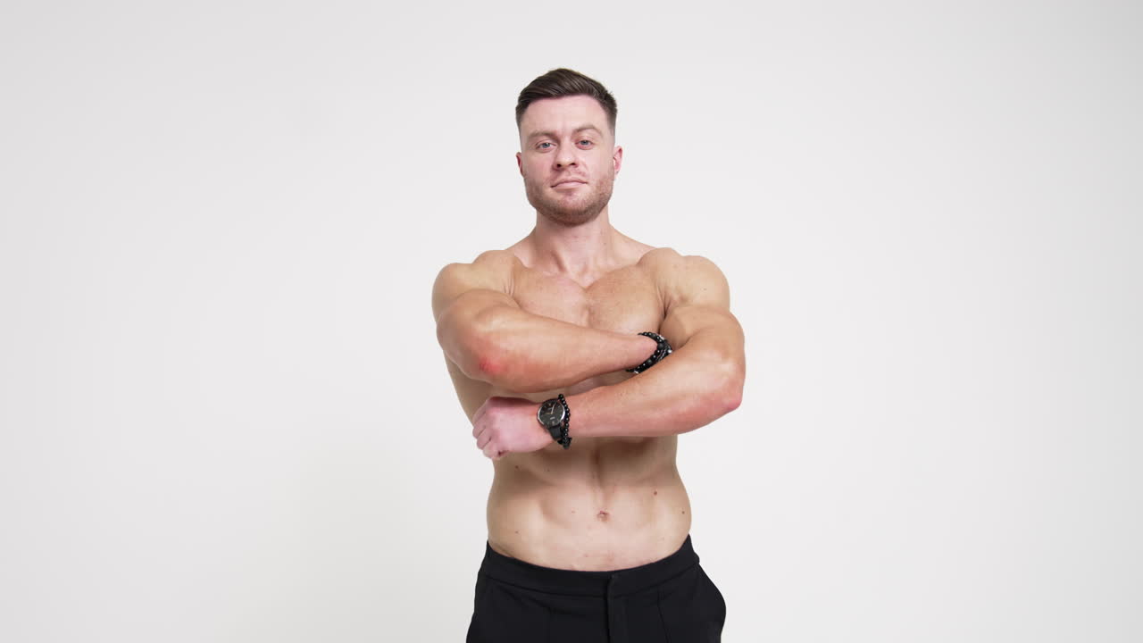 Muscular strong Caucasian male with no shirt in black trousers. Athletic man folding hands on chest looking at camera. White backdrop.
