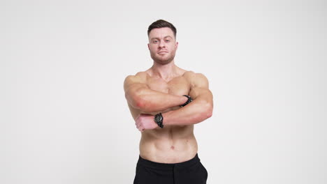 muscular strong caucasian male with no shirt in black trousers. athletic man folding hands on chest looking at camera. white backdrop.