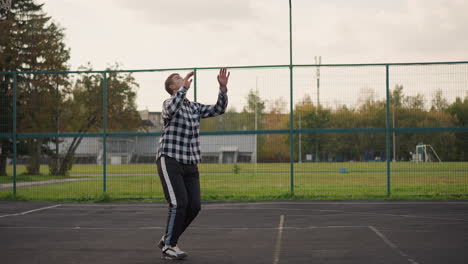 man in plaid shirt on outdoor court tossing volleyball up, preparing to strike with background showing green field and sports equipment, intense sports action and athleticism