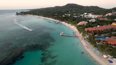 Aerial-view-of-the-coast,-speed-boats,-banana-boat,-green-palms-on-the-sandy-beach-at-sunset