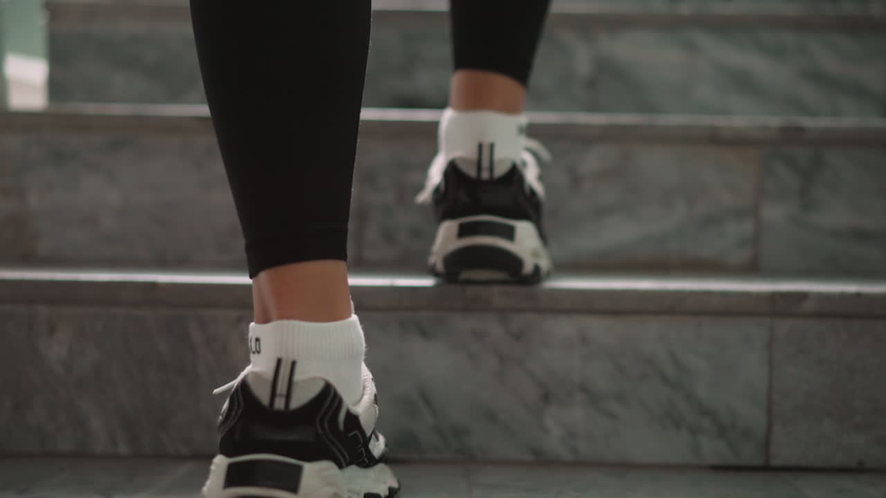 Close view of woman legs in black leggings and white black sneakers climbing marble staircase indoors, careful upward steps with focus on soles and socks, sporty steady motion suggesting fitness