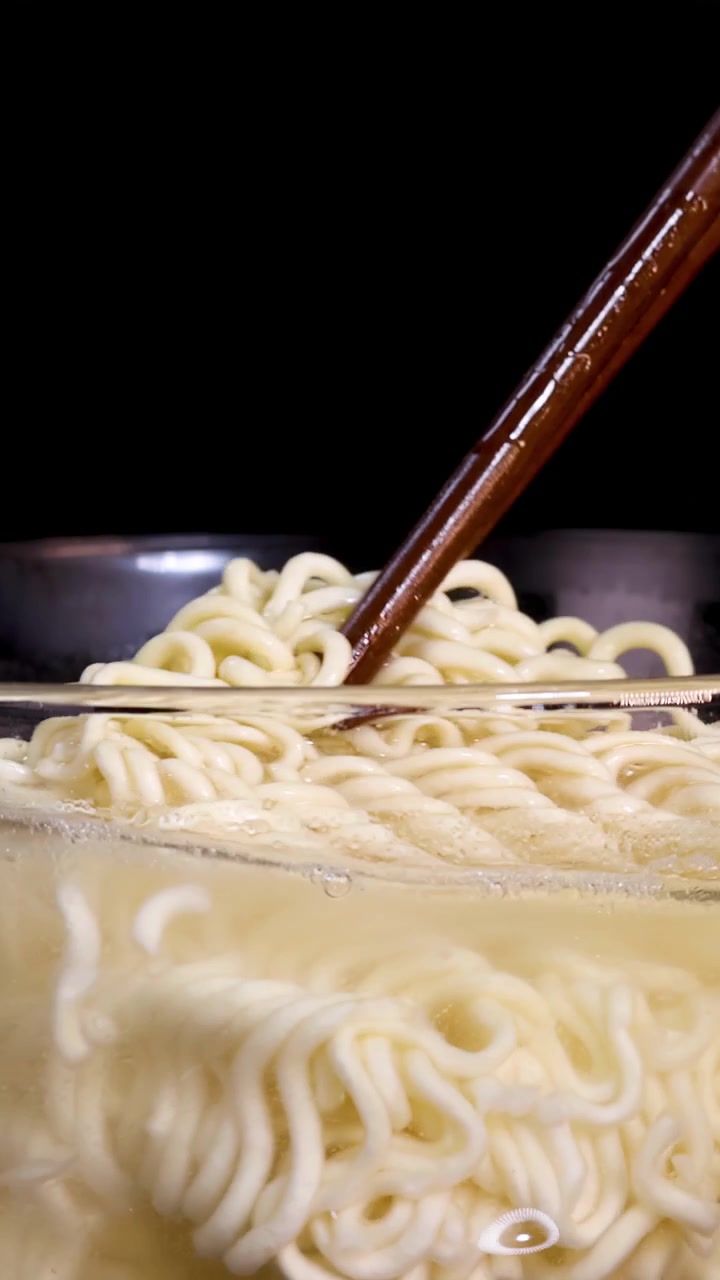 Un paio di bacchette mescola noodles istantanei in una ciotola trasparente di brodo caldo, sotto un'illuminazione drammatica con uno sfondo scuro, ripreso in primo piano.