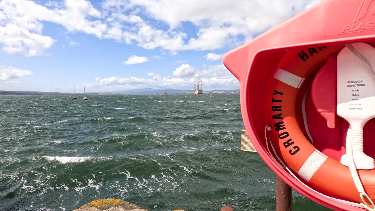 A stationary camera alternates between a close-up of a lifebuoy and a wide view of rough sea with sailing boats, under bright daylight and scattered clouds