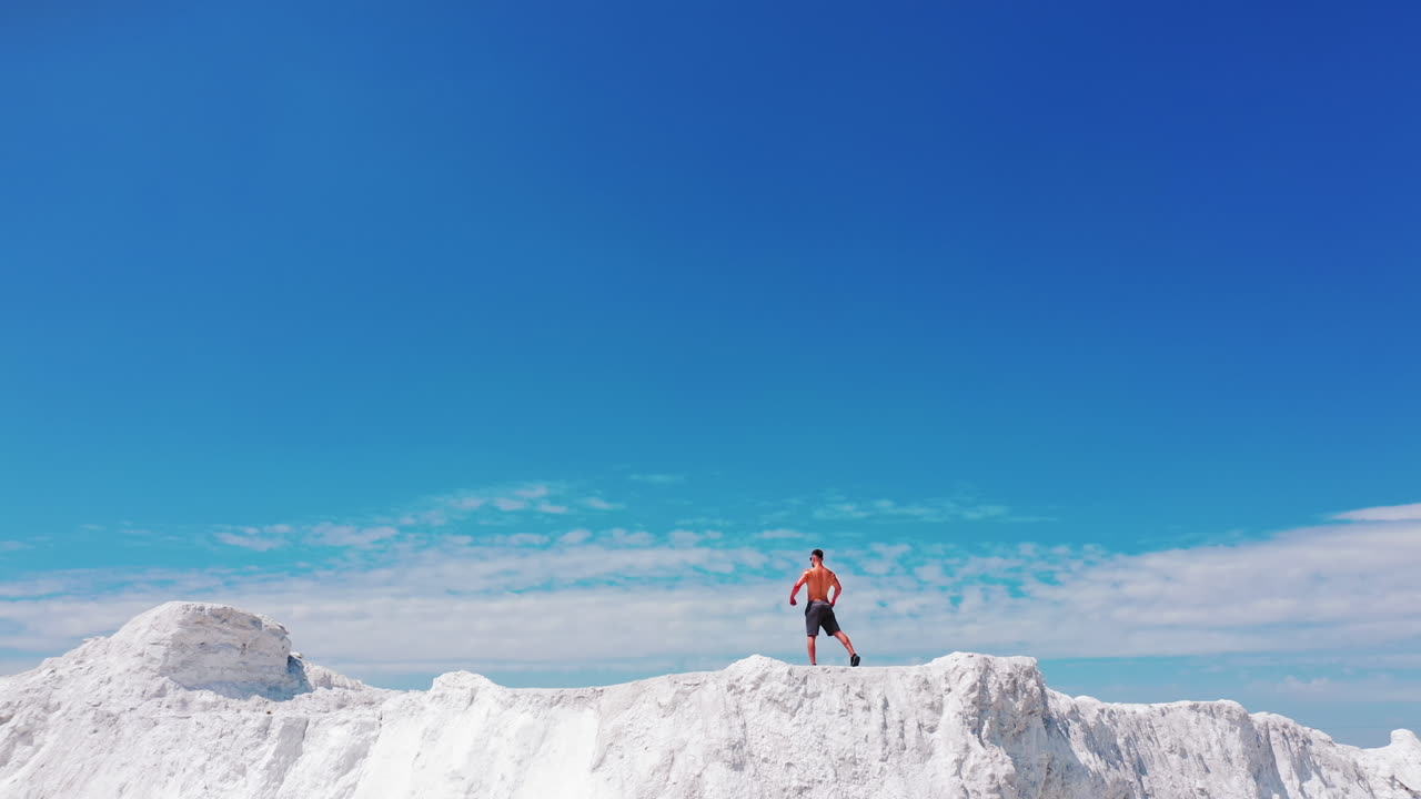 Sportive man in shorts showing his trained body on white hill. Back view of a strong athlete standing in pose on the mountain under blue sky in summer.