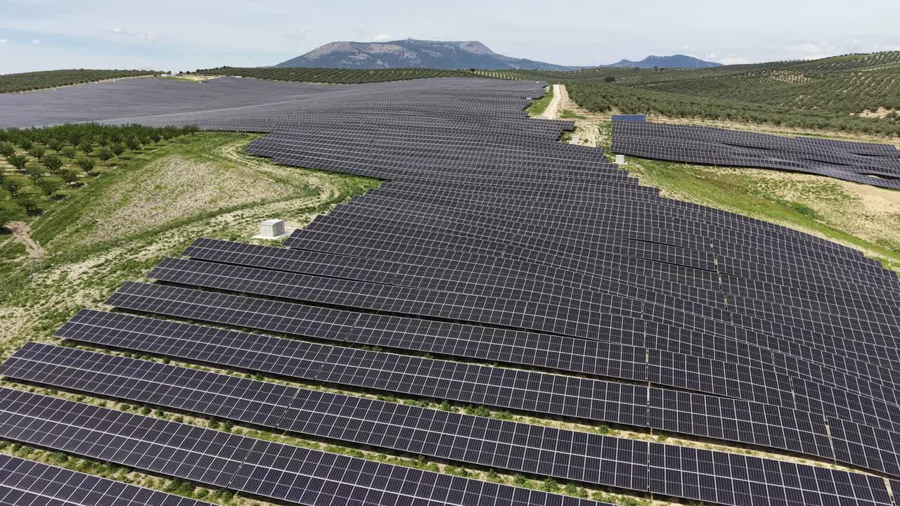 En sporingsdroneoptagelse, der bevæger sig hen over de geometriske mønstre af Pinos Puente solcelleanlægget med bølgende bakker og oliventræer i baggrunden