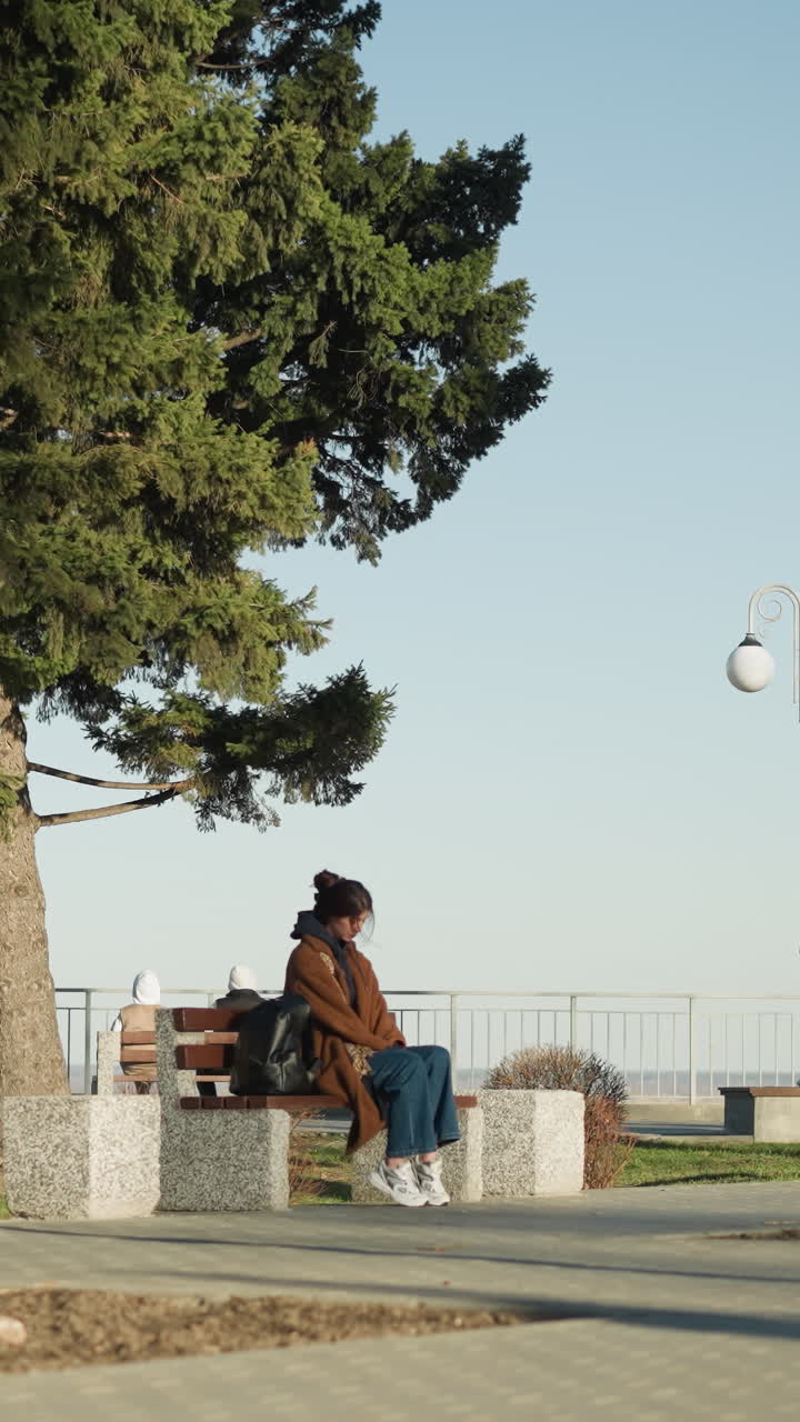 A young woman wearing a brown coat, blue jeans, and white shoes sits alone on a bench in an urban park, her black backpack beside her. She appears moody and deep in thought, with a somber expression