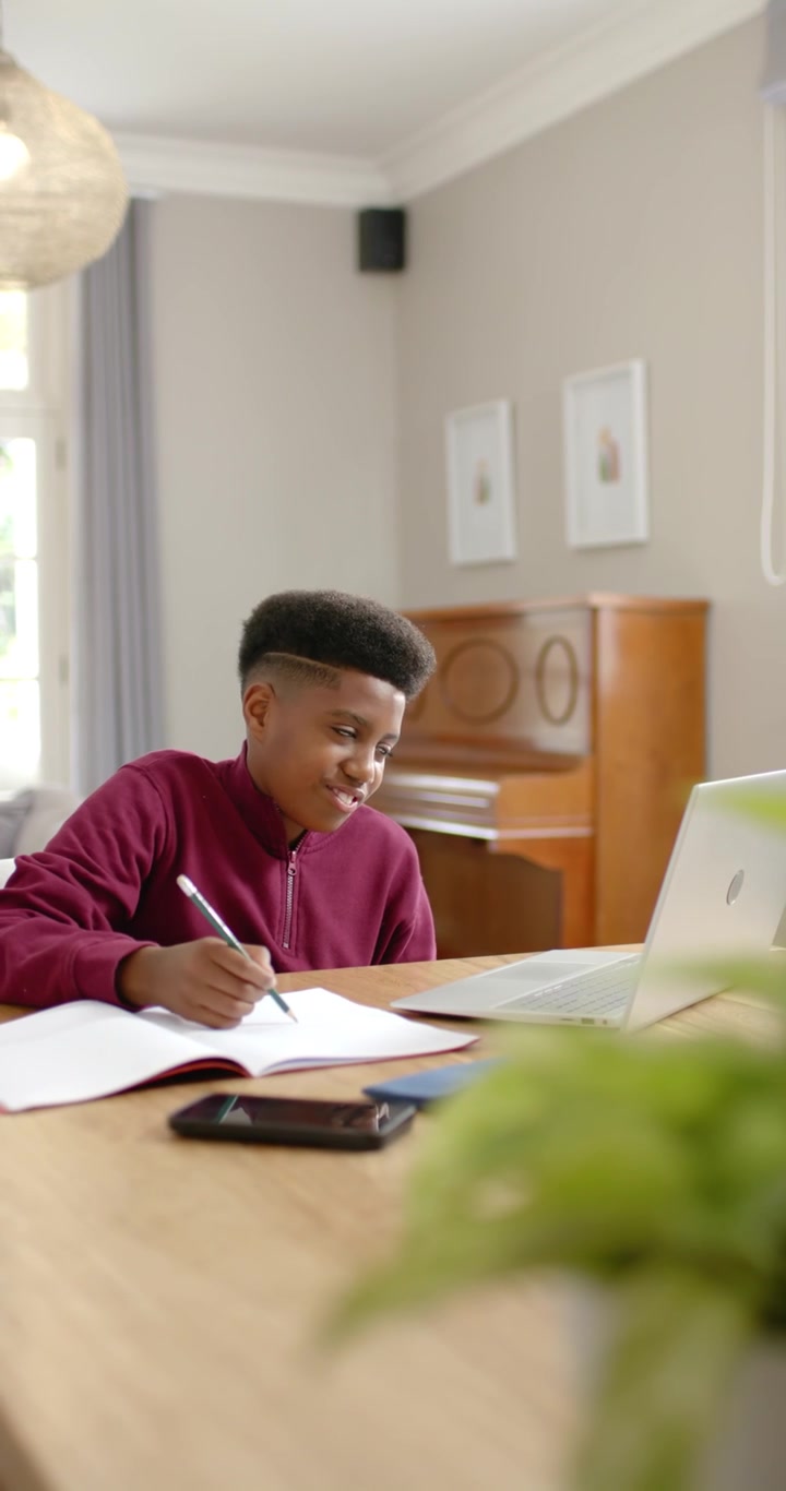 Vertikal video: Ung afrikansk mann sitter hjemme, kikker på laptopen og skriver notater for studier.