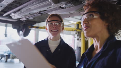 Nachhilfelehrerin-Mit-Studentin,-Die-Im-Kfz-Mechaniker-Kurs-Auf-Einer-Hydraulischen-Rampe-Unter-Das-Auto-Schaut