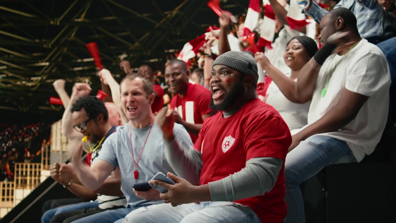 Diverse soccer supporters celebrating in packed stadium stands with red scarves