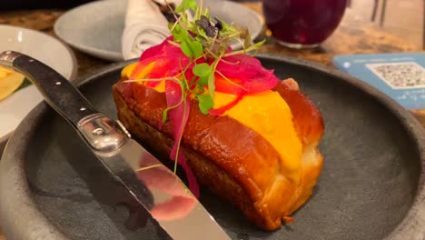 Soft-brioche-bun-filled-with-delicious-meat,-pickled-red-onions,-yellow-sauce-and-microgreens,-delicious-food-on-a-plate,-4K-shot