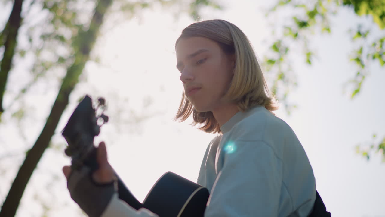 Junger Gitarrist spielt im Freien Akustikgitarre, sonnenbeschienenes, von hinten beleuchtetes Porträt unter belaubten Bäumen, sanftes Klimpern eines Singer-Songwriters, introspektive Stimmung, filmreifes, weiches Bokeh, lässiges Jeanshemd