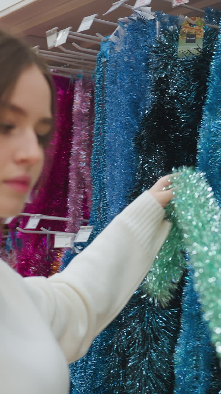 Elegant lady picks shiny green Christmas decoration from shelf in store, carefully observing it, beautiful holiday decorations on display in retail shop with vibrant colors