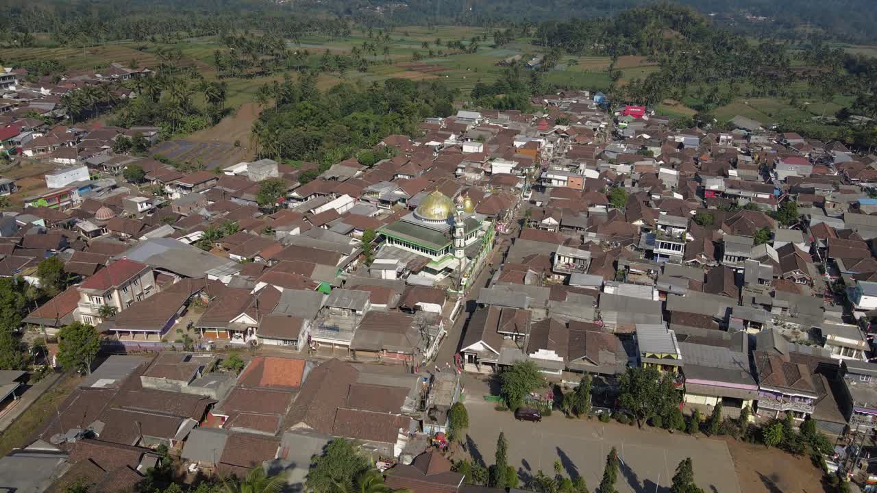 Aerial drone view of Indonesian small town with dense traditional rooftops and central mosque with golden dome, surrounded by tropical landscape and green hills