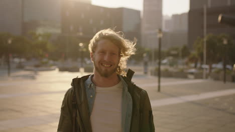 portrait of handsome bearded man looking at camera smiling cheerful enjoying sunset in windy urban city background